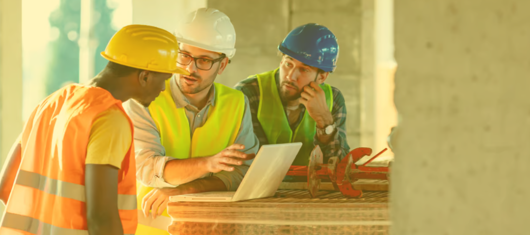 Three construction workers wearing safety vests and hard hats discuss something around a laptop on a worksite, with stacked wooden boards and tools in the background.