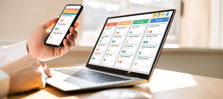 A person holds a smartphone and works on a laptop displaying a digital kanban board with task columns titled To Do, In Progress, Testing, and Done in a bright office setting.