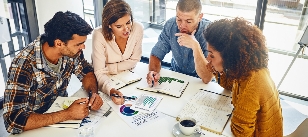 Four people sit around a table in a bright office, discussing charts and graphs. With notebooks and papers in front of them, they appear to be engaged in a collaborative business meeting focused on growing businesses.