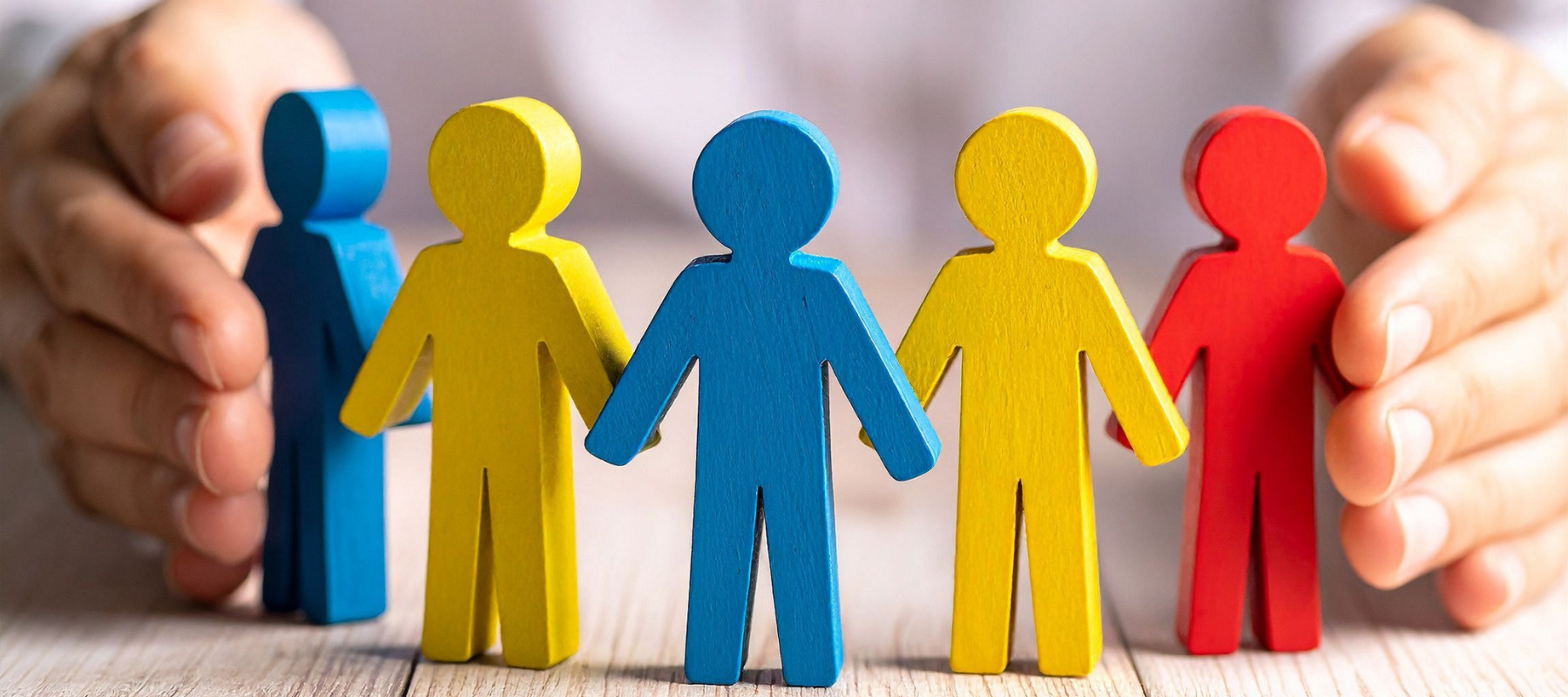Five colorful wooden human figures—two yellow, one blue, one red, and one orange—stand in a row on a wooden surface, with hands curved protectively around them in the background, symbolizing the importance of safety highlighted by WHS Audits.