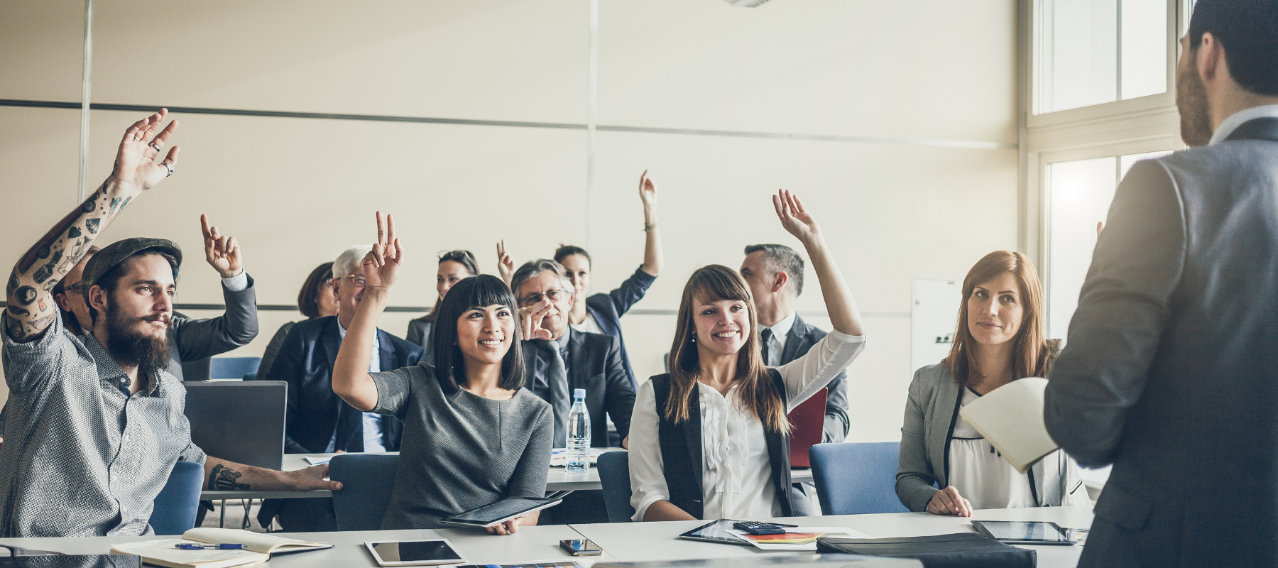 A group of people in business attire sit at desks in a classroom, raising their hands toward a speaker at the front, suggesting participation in a workplace safety seminar or WHS Training session.