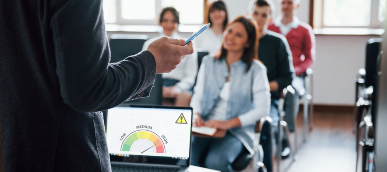 A person gives a presentation to a seated group. In the foreground, a laptop displays an HSR Support gauge labeled low to high with a warning symbol. The audience is attentive and smiling in a bright room.