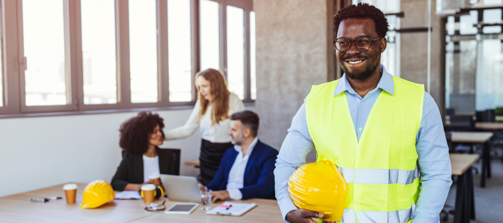 A smiling man in a yellow safety vest, representing HSR Support, holds a hard hat in an office, with three colleagues having a meeting in the background.